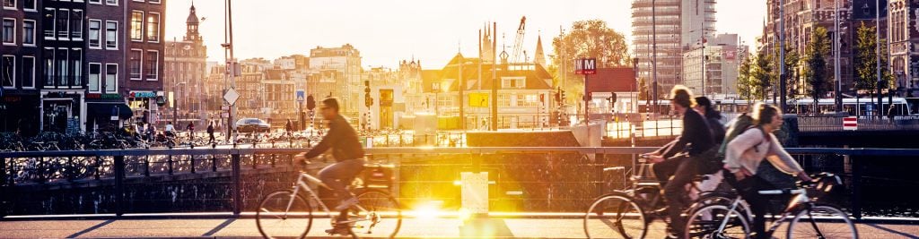Cyclists riding along a canal-side street in Amsterdam at sunset, with historic buildings, bridges, and city infrastructure in the background.