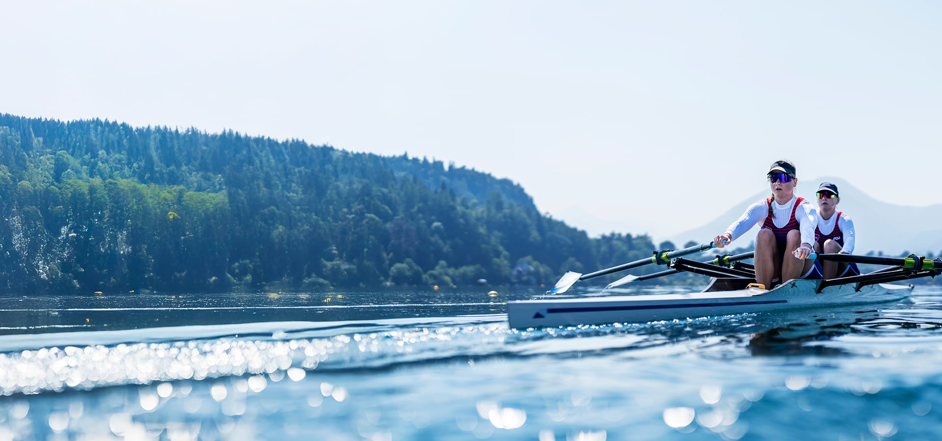 Two rowers in a racing shell glide across a calm lake, creating ripples on the water, with a forested shoreline and distant mountains in the background.